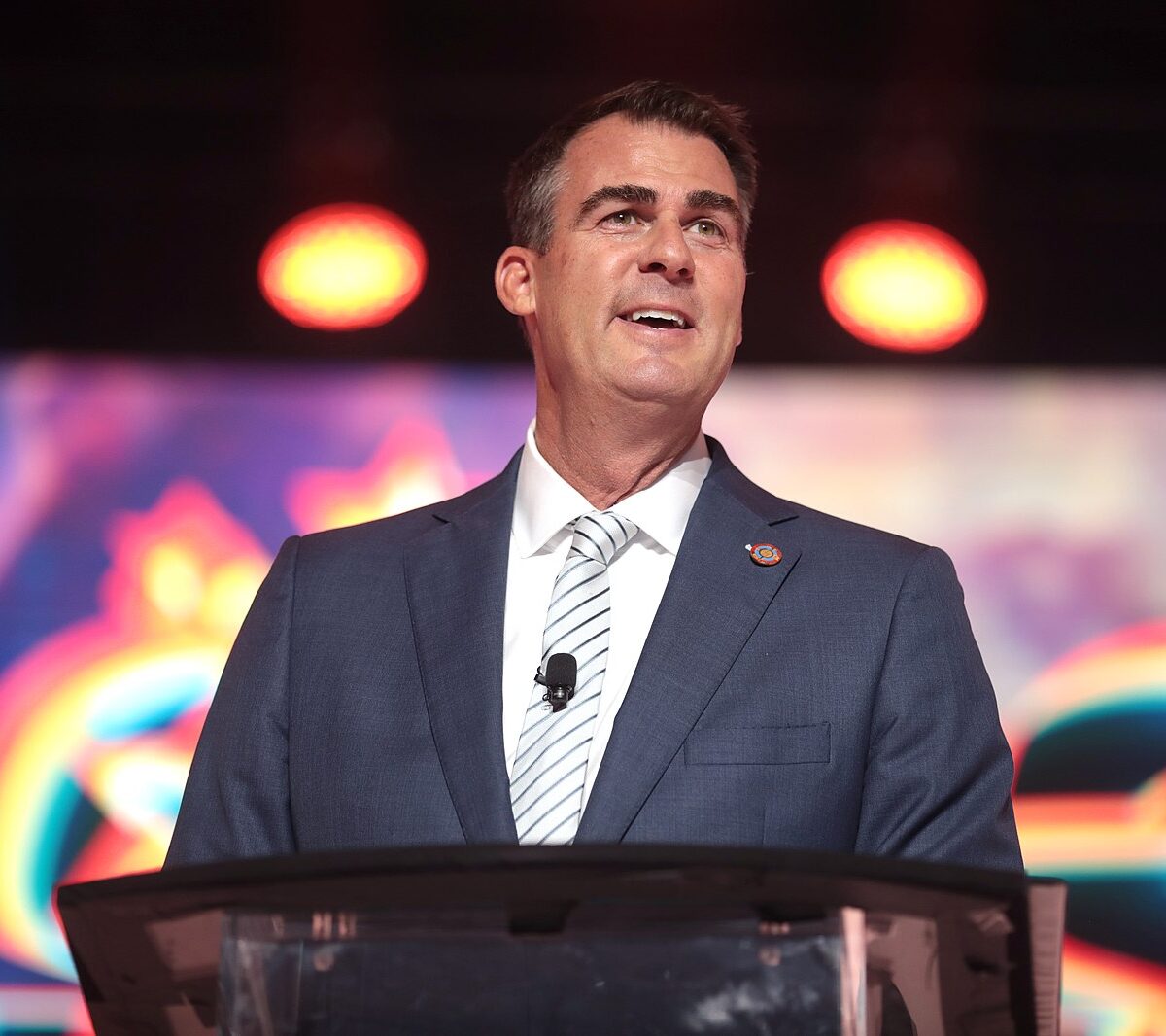 Gov. Stitt Kevin Stitt stands in front of a microphone.